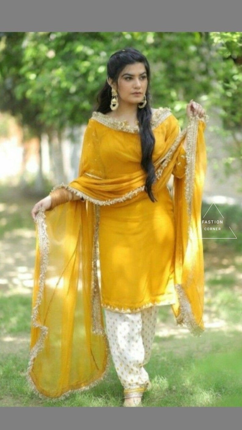 Puede incluir: Una mujer lleva un salwar kameez amarillo con un dupatta blanco y pantalones blancos. El atuendo tiene bordados dorados y un ribete con volantes. La mujer est&aacute; de pie en un campo de hierba.