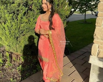 May include: A woman wearing a peach-colored salwar kameez with gold embroidery and a matching dupatta. The dupatta has a gold border and a sheer fabric. The woman is standing on a brick walkway with green trees in the background.