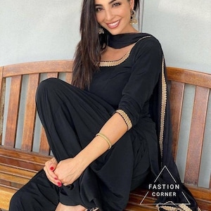 Könnte beinhalten: Ein schwarzer Salwar Kameez mit langen Ärmeln, einem Rundhalsausschnitt mit Goldbesatz und einem passenden Dupatta mit goldenen Bordürendetails. Das Outfit umfasst ein Paar dunkelblaue, bestickte spitze Ballerinas. Text: FASTION CORNER.