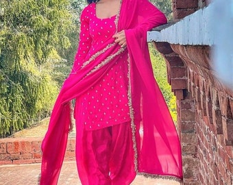 May include: A woman wearing a pink salwar kameez with gold embroidery and a matching pink dupatta. She is standing on a brick wall and looking to the side.