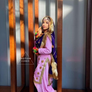 Op de afbeelding: Een vrouw in een roze en paarse outfit met gouden borduurwerk en een paarse dupatta. Ze staat voor een houten muur.