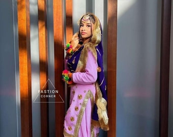 May include: A woman wearing a pink and purple outfit with gold embroidery and a purple dupatta. She is standing in front of a wooden wall.