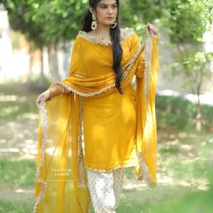 Puede incluir: Una mujer vistiendo un salwar kameez amarillo mostaza con bordados dorados y un dupatta blanco con ribetes dorados. Est&aacute; de pie en un jard&iacute;n con follaje verde en el fondo.