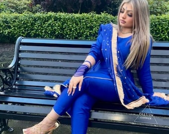 May include: A woman wearing a royal blue pantsuit with gold embroidery and a matching dupatta. She is sitting on a black wooden bench and wearing gold strappy heels. The image has the text "Fashion Corner" in the bottom right corner.