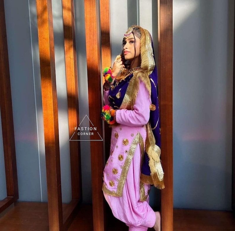 Op de afbeelding: Een vrouw draagt een roze salwar kameez met gouden borduurwerk en een paarse dupatta met gouden rand. De dupatta is over haar hoofd en schouders gedrapeerd. Ze staat voor een houten muur.