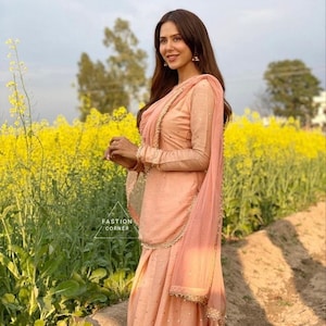 Pu&ograve; includere: Una persona indossa un salwar kameez color pesca, un abito tradizionale del sud asiatico, in un campo di fiori gialli. L'abito comprende una tunica a maniche lunghe, pantaloni abbinati e una sciarpa drappeggiata. L'abito &egrave; adornato con finiture dorate e piccoli abbellimenti. Il testo "FASHION CORNER" &egrave; visibile.
