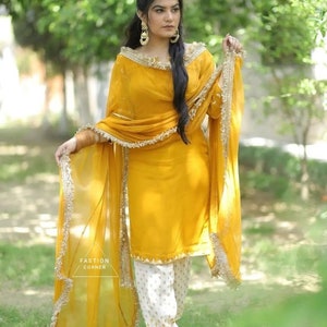 Puede incluir: Una mujer lleva un salwar kameez amarillo mostaza con bordados dorados y un dupatta blanco con ribetes dorados. Est&aacute; de pie en un jard&iacute;n con follaje verde en el fondo.