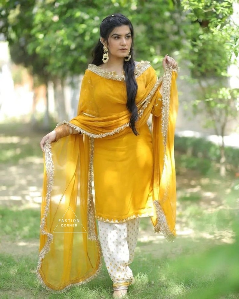 Puede incluir: Una mujer lleva un salwar kameez amarillo mostaza con bordados dorados y un dupatta blanco con ribetes dorados. Est&aacute; de pie en un jard&iacute;n con follaje verde en el fondo.