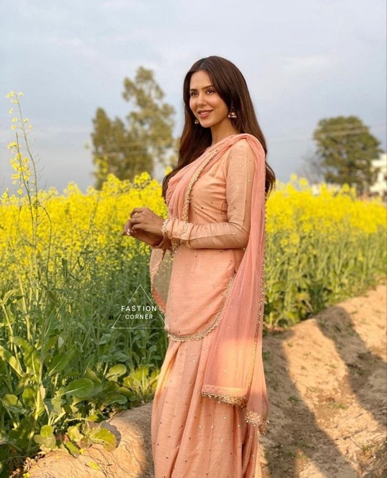 Pu&ograve; includere: Una persona indossa un salwar kameez color pesca, un abito tradizionale del sud asiatico, in un campo di fiori gialli. L'abito comprende una tunica a maniche lunghe, pantaloni abbinati e una sciarpa drappeggiata. Il testo "FASHION CORNER" &egrave; visibile.