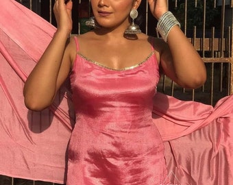 Op de afbeelding: Een vrouw draagt een roze satijnen jurk met een dunne gouden band. Ze draagt grote zilveren oorbellen en een zilveren armband. De jurk is strak en heeft een ronde halslijn. De vrouw staat voor een metalen hek.