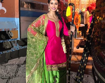 May include: A woman wearing a pink and green salwar kameez with gold embroidery. She is holding a black and gold walking stick with a yellow tassel.
