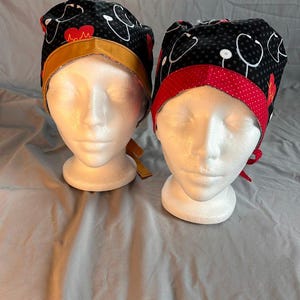 May include: Two scrub caps on mannequin heads. One cap has a black background with white stethoscopes and red hearts. The other cap has a black background with white stethoscopes and red polka dots. Both caps have a contrasting trim.
