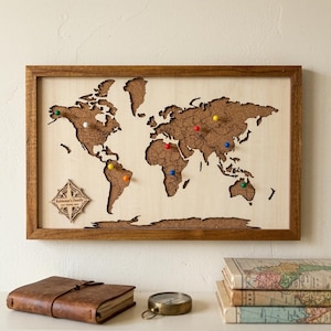 May include: A rectangular wooden framed world map with a cork inlay featuring laser-cut continents. The map includes a compass rose with the text 'Robinson's Family OUR TRAVEL MAP' and several colorful push pins placed across various regions.