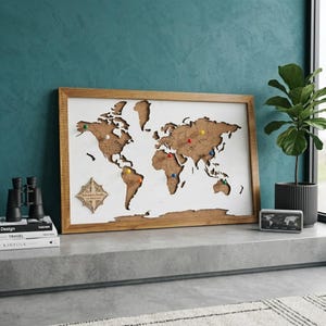 Puede incluir: Un mapa del mundo rectangular enmarcado con fondo blanco y continentes de corcho. El mapa presenta una rosa de los vientos de madera en la esquina inferior izquierda con el texto "Bohomme's Family THE WORLD MAP". Varias chinchetas de colores.