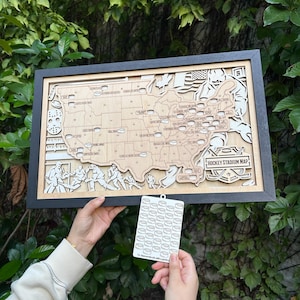 May include: A framed wooden map of the United States, laser-cut to show hockey stadium locations. The map is in a black frame and has the text "HOCKEY STADIUM MAP". A small tag with hockey pucks is held in front of the map.