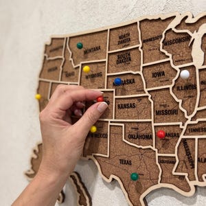May include: A wall-mounted cork map of the United States featuring laser-cut wooden borders for each state. The map includes black printed state names and major city labels. Several colourful push pins are inserted into various locations.