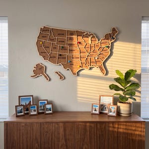 May include: A large, wooden, laser-cut map of the United States of America mounted on a wall, featuring individual state segments, engraved state names, and several colourful push pins inserted into various locations across the map.