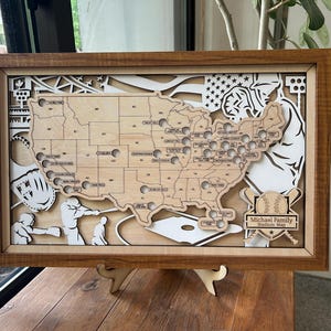 May include: Wooden framed baseball stadium map of the United States. The map is laser cut with a light wood finish, featuring stadium locations and baseball-themed cutouts. The frame is a dark brown color. The text "Michael Family Stadium Map" is visible.