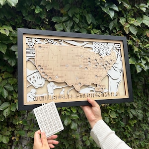 May include: A framed wooden baseball stadium map of the United States, with stadium locations marked. The map is layered with a black frame and includes baseball-themed illustrations. A small tag with baseball stickers is held below.