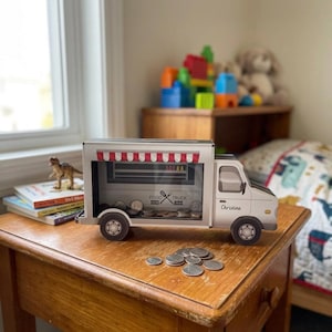 May include: A white food truck-shaped bank with a red and white striped awning. The bank has a clear window showing coins inside. The text "FOOD TRUCK" and "Christina" are visible on the bank. The bank is sitting on a wooden table.