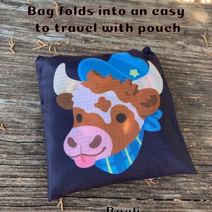 May include: A black reusable shopping bag with a cartoon cow wearing a blue hat and a blue and green scarf. The bag folds into an easy to travel with pouch. The text "Bag folds into an easy to travel with pouch" is printed on the bag. The text "Back" is printed on the wooden surface below the bag.