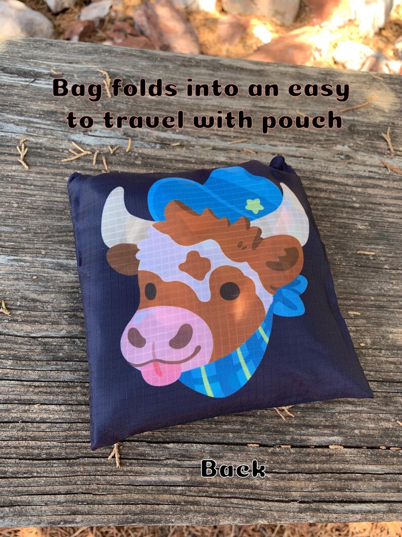 May include: A black reusable shopping bag with a cartoon cow wearing a blue hat and a blue and green scarf. The bag folds into an easy to travel with pouch. The text "Bag folds into an easy to travel with pouch" is printed on the bag. The text "Back" is printed on the wooden surface below the bag.