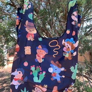 May include: A black reusable shopping bag with a colorful pattern of cartoon animals wearing cowboy hats and holding various items. The animals include a cat, a cow, a bird, and a llama. The bag also features cacti, oranges, and a lasso.