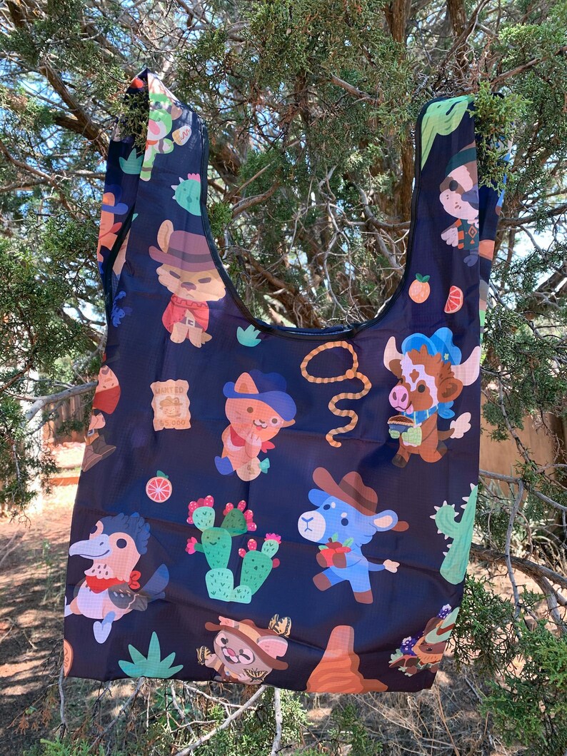 May include: A black reusable shopping bag with a colorful pattern of cartoon animals wearing cowboy hats and holding various items. The animals include a cat, a cow, a bird, and a llama. The bag also features cacti, oranges, and a lasso.