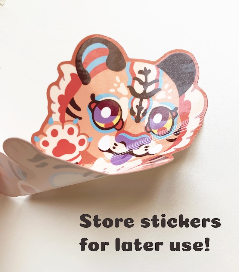 May include: A colorful sticker featuring a cartoon tiger with a pink, blue, and yellow design. The tiger has large, purple eyes and a white paw print on its front leg. The sticker is partially peeled off of a white surface.