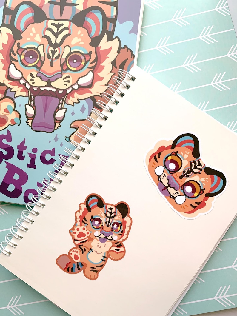 May include: Two colorful tiger stickers on a white notebook. The stickers feature a tiger with orange, yellow, and blue stripes. The notebook has a spiral binding.
