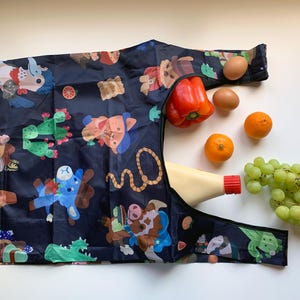 May include: A black reusable shopping bag with a colorful cartoon print of animals and plants. The bag is filled with groceries, including a red pepper, oranges, grapes, and a bottle of mayonnaise.
