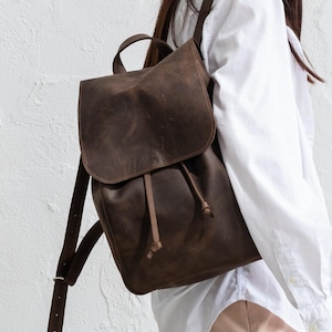 Peut inclure: Un sac à dos en cuir marron foncé doté d'un rabat repliable avec fermeture à cordon, d'une poignée supérieure et de bretelles réglables. Le sac présente une finition mate et texturée et est porté au dos avec les lanières pendantes.
