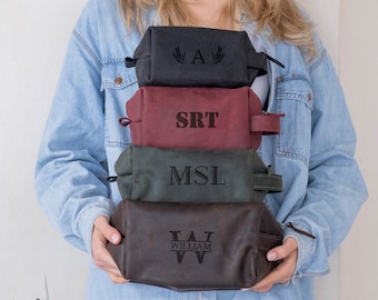 Puede incluir: Una pila de cuatro neceseres rectangulares en negro, burdeos, verde oliva y marrón oscuro. Cada bolsa cuenta con un asa lateral, cierre de cremallera y un monograma o nombre grabado, incluyendo las letras A, SRT, MSL y WILLIAM.
