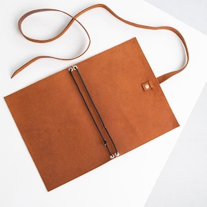 May include: A brown leather journal with a long strap and a black elastic closure. The journal is open, revealing the interior pages. The strap is made of the same brown leather as the journal.