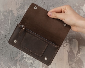 May include: A dark brown leather wallet featuring a fold-over flap with two silver-toned metal snap buttons, a horizontal zippered compartment, and a rectangular card slot pocket on the interior surface, constructed with visible stitching.