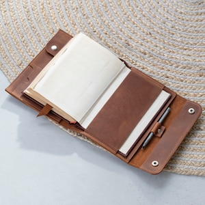 May include: An open brown leather journal cover featuring a wrap-around design with two silver metal snap closures. The interior includes a cream-colored notebook, a leather bookmark, and a dedicated pen loop holding a silver metallic pen.
