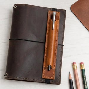Puede incluir: Una funda de diario de cuero marrón oscuro con un portalápices vertical de cuero color canela claro sujeto por una banda elástica negra. La funda se asegura con un cordón elástico negro fino e incluye ojales metálicos en las esquinas.