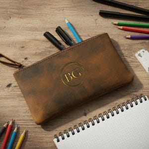 Può includere: Un astuccio in pelle marrone con cerniera e un emblema circolare dorato con le lettere BG impresse al centro. L'astuccio è dotato di una linguetta in pelle ed è circondato da varie matite colorate, penne nere, una gomma bianca e un quaderno a spirale.