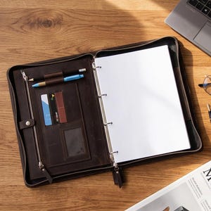 May include: A dark brown leather zippered binder featuring a metal ring mechanism, a vertical zippered pocket, two card slots, a clear ID window, and two pen loops holding a black pencil and a blue pen, containing blank white paper sheets.