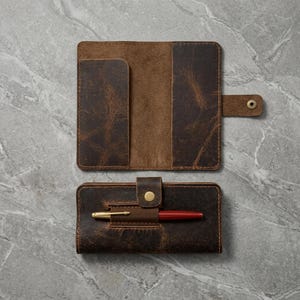 May include: A dark brown leather notebook cover with a distressed finish, featuring a snap closure tab and an integrated elastic pen loop. The cover is shown in both an open position and a closed position with a red and gold pen inserted.