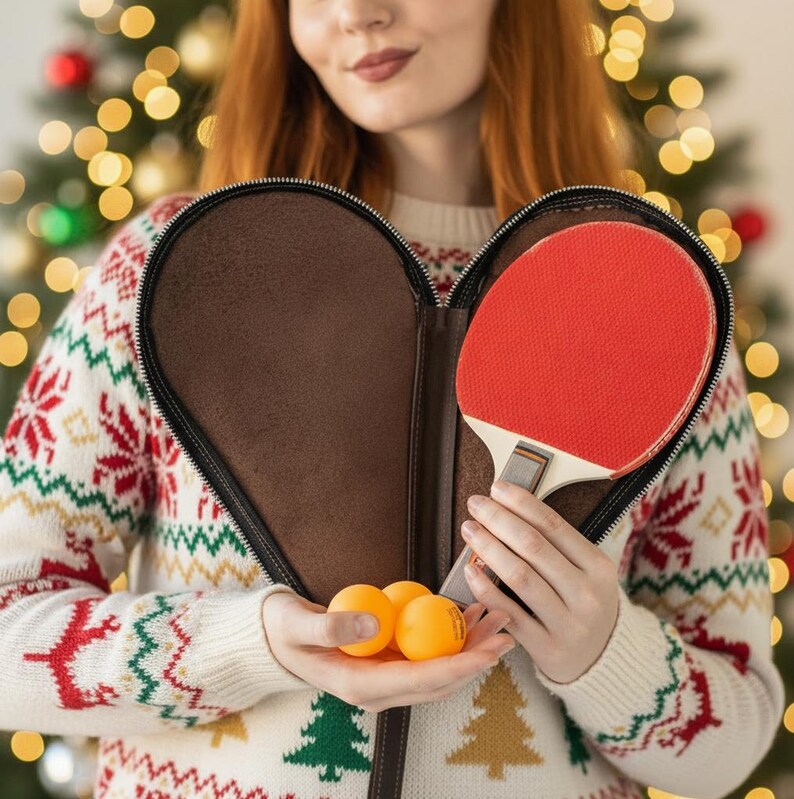 May include: A red ping pong paddle with a white handle is held in a brown case. Three orange ping pong balls are held in the other hand. The person is wearing a white sweater with a Christmas pattern.