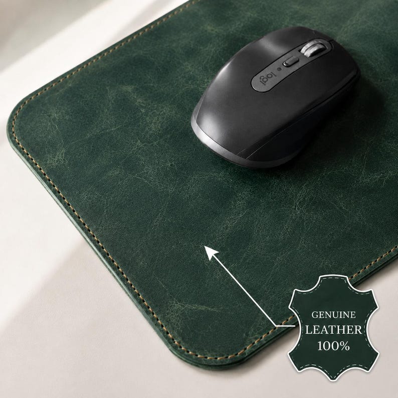 K&ouml;nnte beinhalten: Ein dunkelgr&uuml;nes, rechteckiges Mauspad aus Leder mit abgerundeten Ecken und sichtbaren N&auml;hten an den R&auml;ndern. Eine schwarze kabellose Computermaus liegt auf der Oberfl&auml;che. Ein kleines Emblem zeigt den Text GENUINE LEATHER 100%.