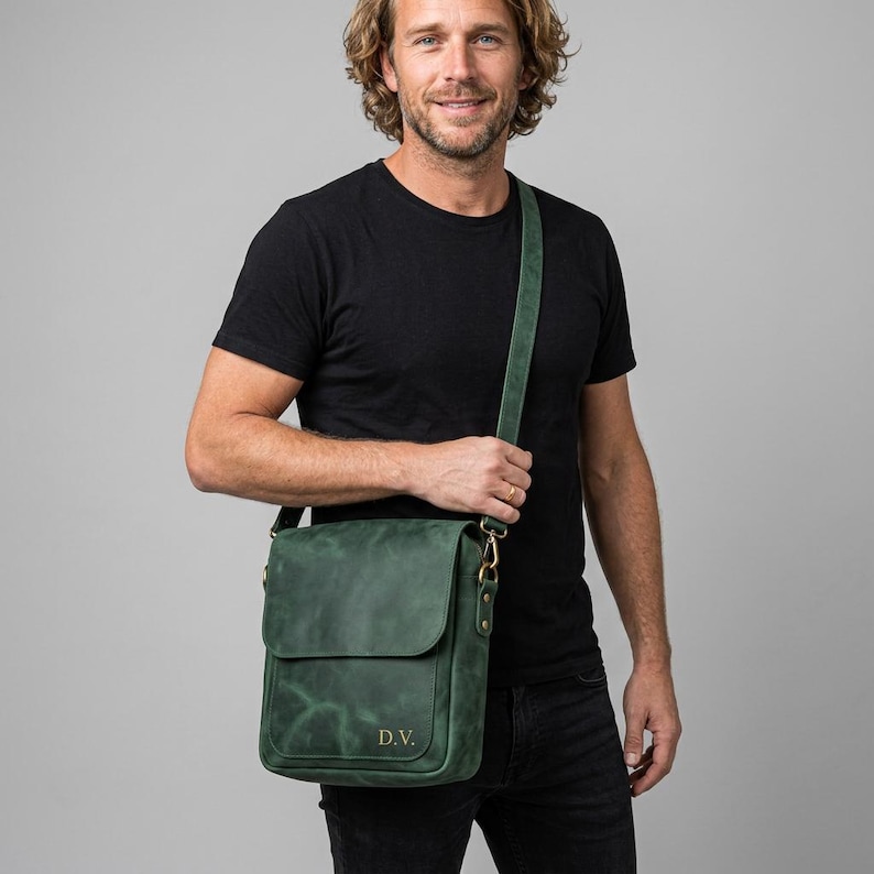 May include: A dark green leather messenger bag with a flap closure and an adjustable shoulder strap. The front of the bag features the embossed initials D.V. in a gold-colored font. The bag has a matte finish and gold-toned metal hardware.