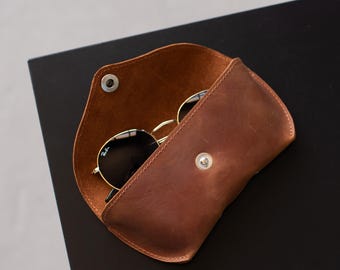 May include: A brown leather glasses case with a snap button closure, containing a pair of black-rimmed sunglasses with gold-tone metal frames. The interior of the case is visible, showing the smooth texture of the leather and the metal snap hardware.