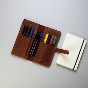 May include: A brown leather pen roll with multiple slots containing black, blue, yellow, red, and green pens, featuring a strap closure and the embossed letters BM, positioned next to a closed white notebook with a black elastic band.