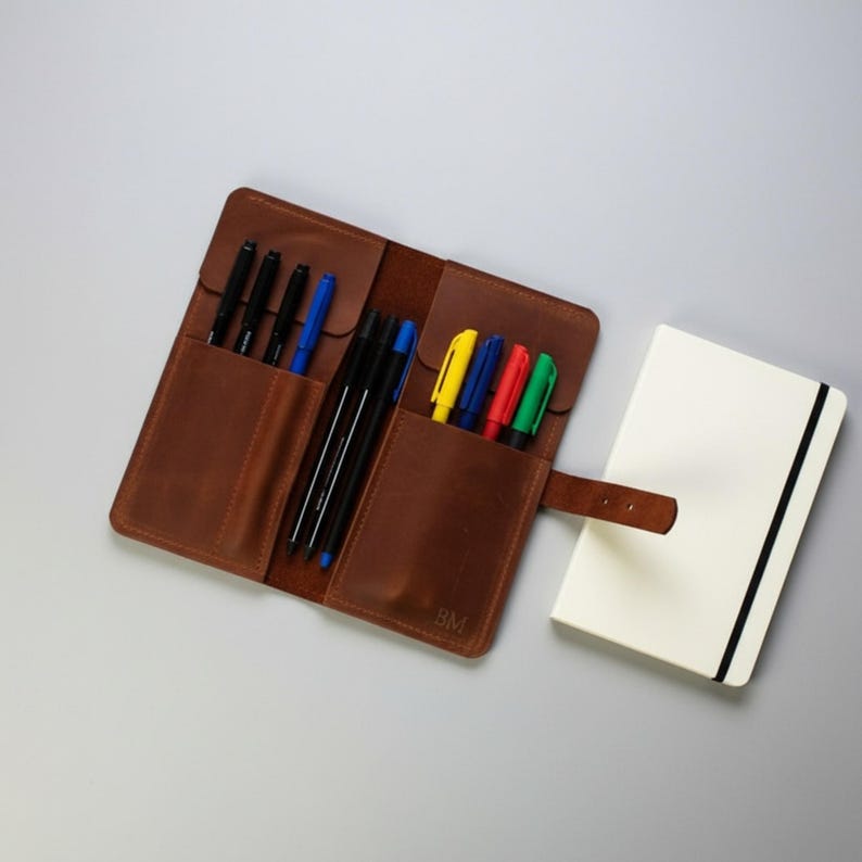 May include: A brown leather pen roll with multiple slots containing black, blue, yellow, red, and green pens, featuring a strap closure and the embossed letters BM, positioned next to a closed white notebook with a black elastic band.