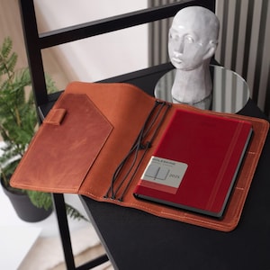 Puede incluir: Una funda de cuero color canela para cuaderno con diseño abierto, bolsillos internos y bandas elásticas negras que sujetan una agenda Moleskine 2025 roja. La funda tiene costuras visibles en los bordes y un portalápices en el panel izquierdo.