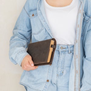 May include: A dark brown leather book cover with visible stitching along the edges, featuring a wrap-around design and a small side strap closure. The cover is shown holding a book with light-colored pages, providing a protective sleeve.