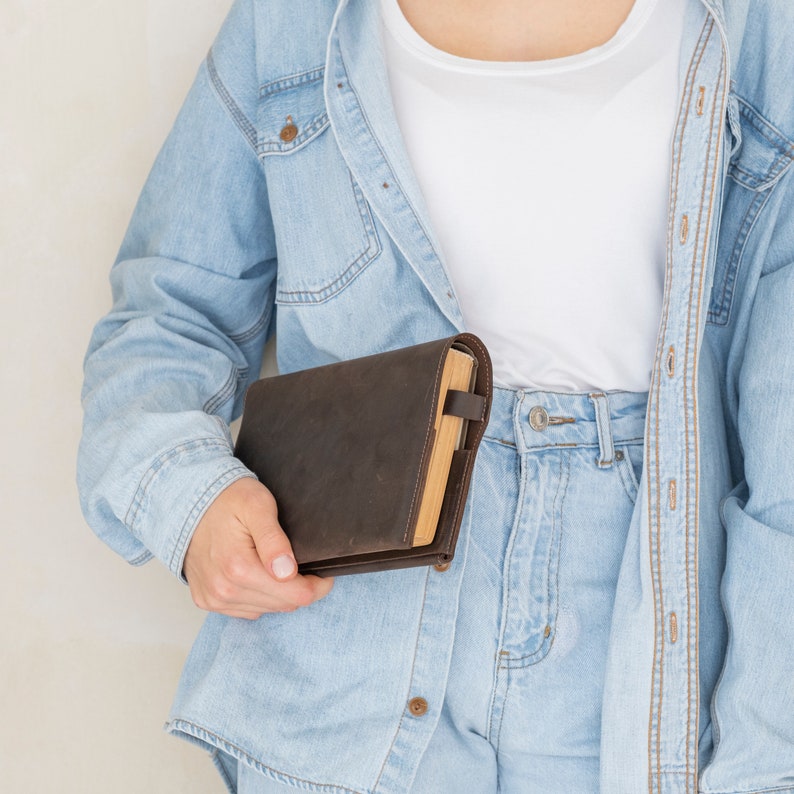 May include: A dark brown leather book cover with visible stitching along the edges, featuring a wrap-around design and a small side strap closure. The cover is shown holding a book with light-colored pages, providing a protective sleeve.