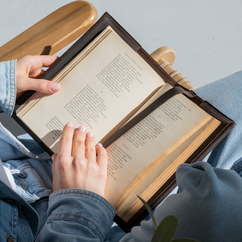 May include: A dark brown leather book cover holding an open book with cream-colored pages featuring printed Russian text. The cover has a smooth texture and stitched edges, providing a protective sleeve for the open volume of literature.